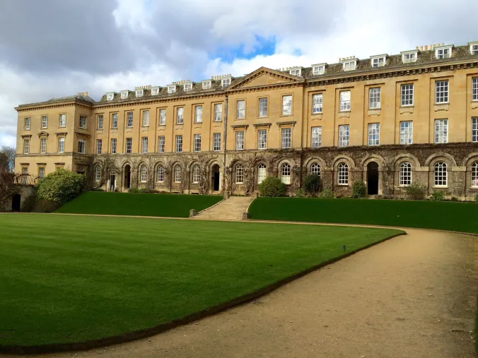 Large stone institutional building with arched walkways facing a wide lawn, under partly cloudy sky, muted earth tones